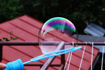 Naklejka premium A bubble attaching on bubble blower use as decoration. On the top of bubble is green and purple filter and in the background there are cords, tree and piece of roof