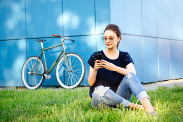 Portrait of young cute girl with smartphone, browsing something or chatting while sitting on grass, after riding a bike. Leisure time.