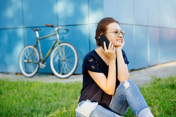 Outdoors photo of cheerful attractive girl, having pleasant phone conversation, resting on grass after riding a bike. Outdoors. Leisure concept.