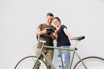 Smiling photographer and happy girl checking photos after photo shooting, standing with retro bike over white wall background. Outdoors.
