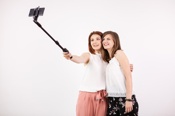 Two female best friends having a lot of fun while taking a selfie with a selfie stick over a white wall in studio