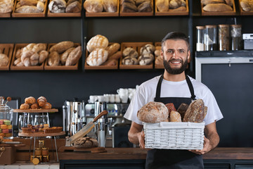 Male baker holding wicker basket with fresh bread in shop - obrazy, fototapety, plakaty