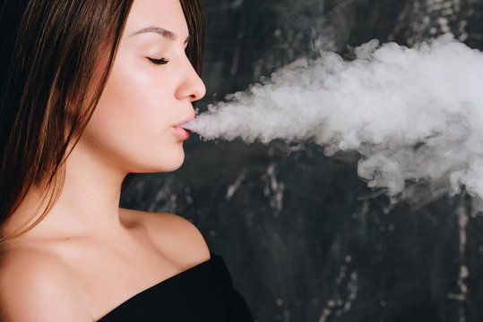 Side view of cute trendy girl exhaling big clouds of smoke from e-cigarette, standing over dark background, indoors.