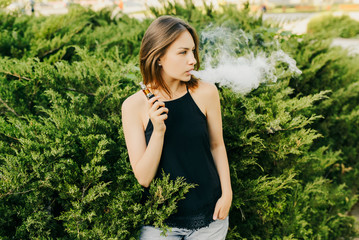 Trendy girl exhaling a cloud of vapor while smoking an electronic cigarette, standing near the bushes, looking away and keeping the hand in the pocket. Outdoors.