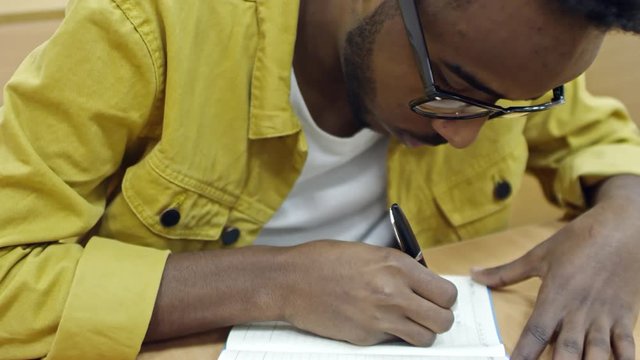 Close up shot of young black man leaning over copybook when taking notes during lecture at university