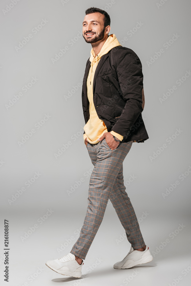 Sticker smiling stylish male model in autumn outfit posing with hands in pockets on grey background