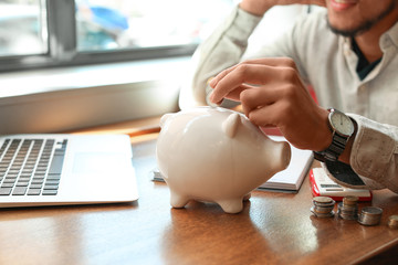 African-American man putting coins into piggy bank on table. Savings concept