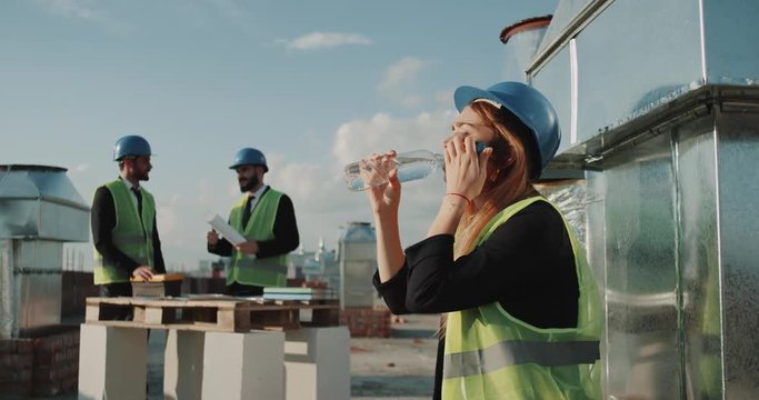 Young woman worker from construction site in hot work day have a little break drinking water , background builders have a conversation.