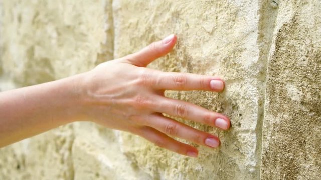 Woman sliding hand against old ancient stone wall in slow motion. Female hand touching hard rough surface of rock on sunny summer day