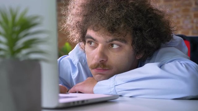 Bored nerdy boss with curly hair and mustache lying on table and working on laptop, modern office background