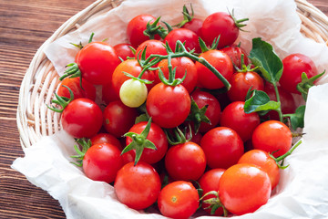 Small red cherry tomatoes spill out of a wicker basket on an old wooden table in rustic style, selective focus