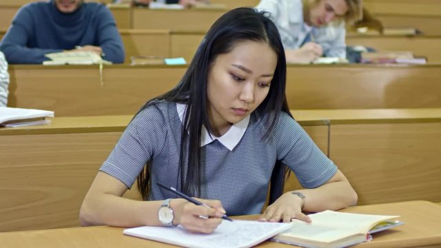 Medium shot of diligent Asian woman sitting at desk in lecture hall and using textbook when writing in notebook, other students sitting in the background, tracking shot