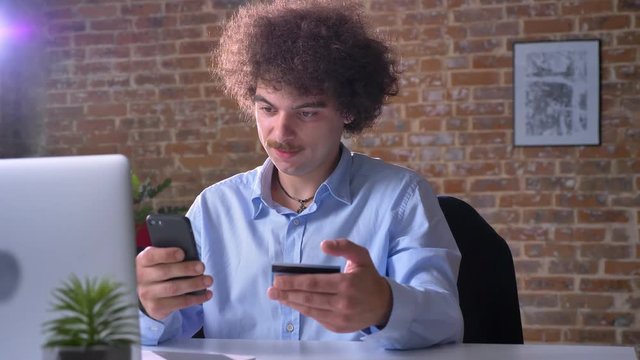 Funny nerdy office worker paying bills in phone with credit card, sitting and shopping in modern office, indoors