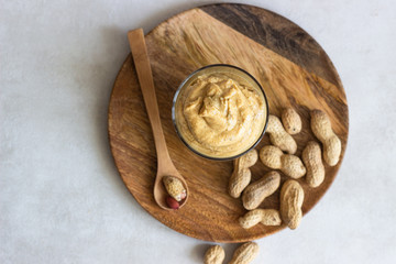 Creamy peanut butter in a glass jar and wooden spoon on light grey background. Copy space.