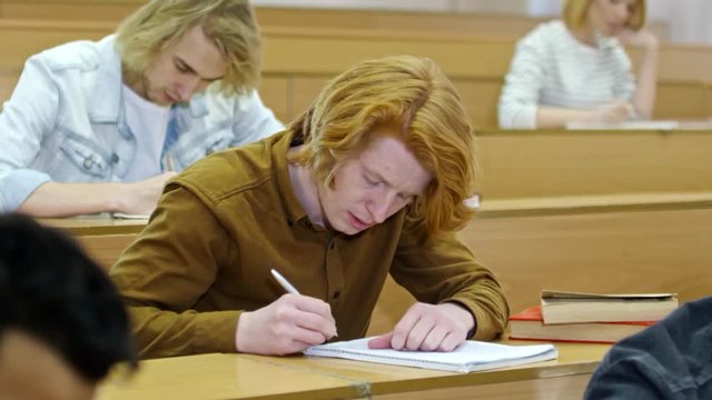Medium shot of ginger haired young man writing in notebook when doing task during lecture at university