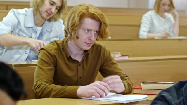 Medium shot of young ginger haired man receiving cheat sheet from group mate when taking test at university