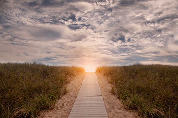 A wooden path leading among the sands and grass on the beach of the Antaltiechi coast. Road going up into the clouds. USA. Maine.
