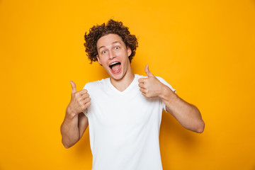 Handsome happy man 20s with brown curly hair shouting and showing thumbs up on camera, isolated...