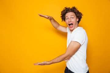 Young excited man 20s with brown curly hair gesturing aside and holding big copyspace in hands,...