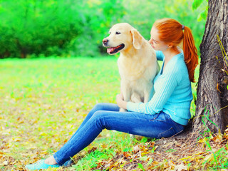 Happy owner woman with Golden Retriever dog together is sitting near a tree in the park