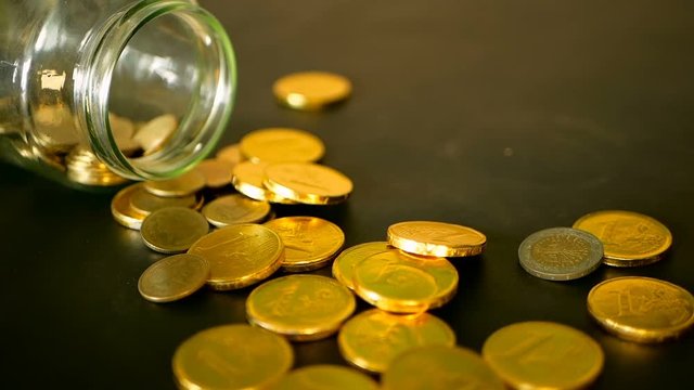 Yellow coins fell out from jar. Symbol of investing, keeping money concept. Collecting cash conis in glass tin as moneybox. Close-up still life with gold coins on black table and rotating penny.