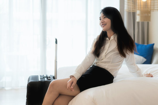 Smiling Asian business woman sitting on bed in hotel room. Business travel concept..