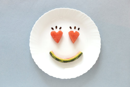 Smiling faces on a white plate on blue pastel background. Heart shape from watermelon. Smiling symbol. Concept enjoy eating. Summer time