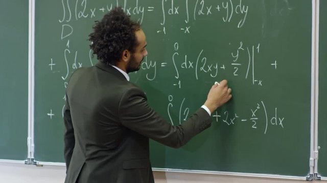 Medium shot of middle-aged teacher of mathematics standing at blackboard with equations written on it and explaining topic to students during lecture at university