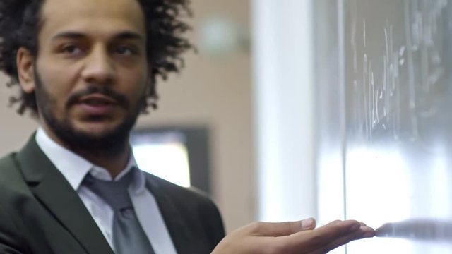 Handheld shot of middle aged university professor pointing at blackboard when asking question to student during lecture