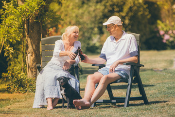 A retired happy couple is sitting in the garden drinking beer with their little black dog.