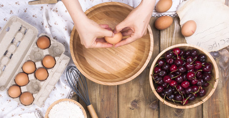 Female Hands break the egg into a wooden bowl. Preparation of dough for a pie with a cherry. Summer homemade pastries. Top view. Copy space.