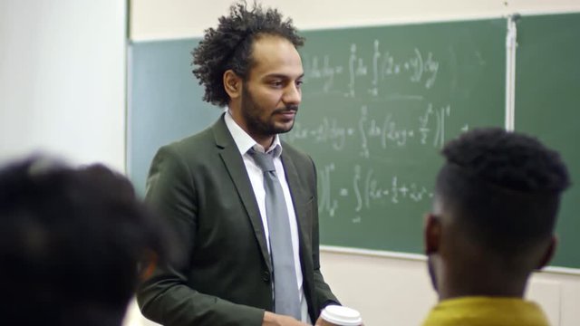 Medium shot of smiling middle eastern man holding takeaway coffee cup and talking to group of students before mathematics lesson, handheld