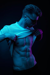 Neon light portrait of sexy muscular man in yellow glasses, pulling up t-shirt, showing abs