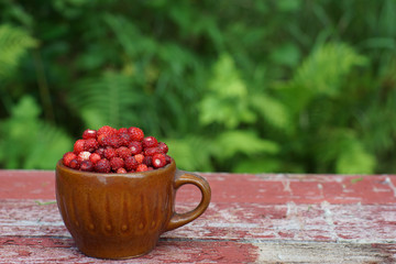 Rustic style mug filled with red strawberries, stands on shabby, faded boards in the garden in...