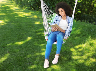 Beautiful young African-American woman reading book in hammock outdoors