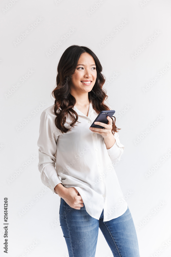 Poster Photo of happy asian woman with long dark hair holding black mobile phone and looking aside, isolated over white background in studio