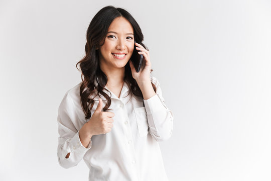 Photo of joyful asian woman with long dark hair holding and speaking on smartphone, isolated over white background in studio