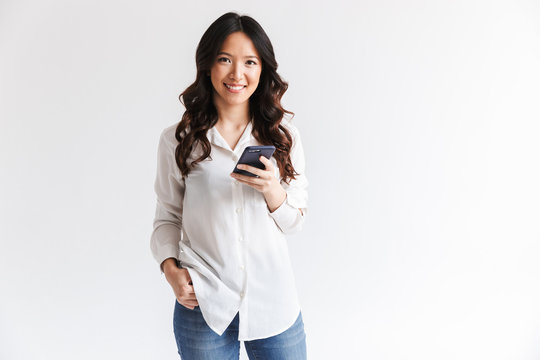 Photo of smiling charming asian woman with long dark hair holding black cell phone, isolated over white background in studio