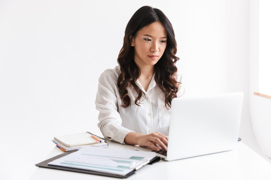 Photo of young asian woman with long dark hair sitting at table and working with documents and laptop, isolated over white background in studio
