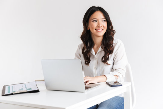 Photo of charming chinese businesswoman with long dark hair sitting at table and working with documents and laptop, isolated over white background in studio