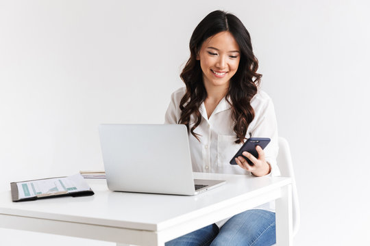 Photo of young asian businesswoman with long dark hair sitting at table and holding smartphone while working with laptop, isolated over white background in studio