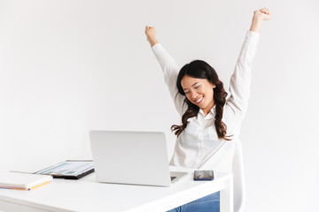 Photo of successful chinese businesslike woman with long dark hair smiling with raised arms while working with documents and laptop, isolated over white background