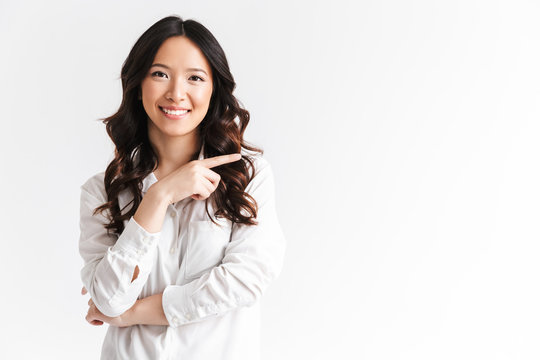 Joyful asian woman with long dark hair looking at camera with beautiful smile and pointing finger aside at copyspace text or product, isolated over white background in studio