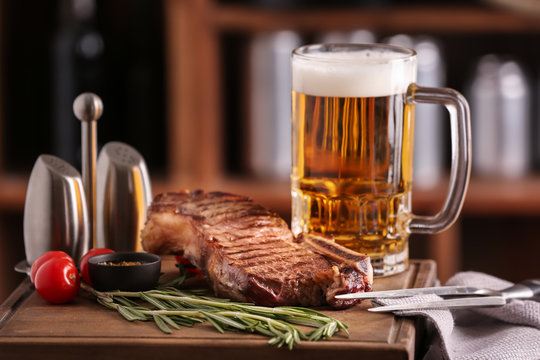 Mug of delicious beer with grilled steak on wooden board