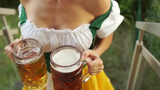 Oktoberfest waitress carries two large glasses of beer. The girl walks barefoot on the grass and climbs the stairs. Bavarian traditional costume, sexy decollete