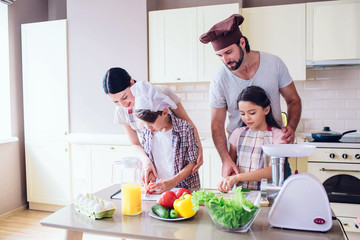 Family is standing in kitchen and cooking. Guy helps girl to cut cucumber. Woman stands behind her son and look at how he cuts tomato with knife.