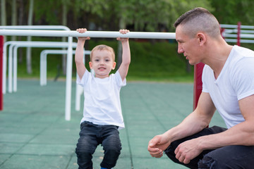 Fototapeta na wymiar A strong athletic father insures the boy on sports equipment and teaches him to pull himself up on the horizontal bar in the summer park. Strong athletic father teaches his son to catch up on the bar.