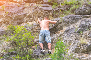 Young man climber in jeans shirts and bare torso climbs on rocks to the top in mountains of Altai without equipment and insurance