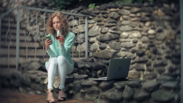 Woman with a laptop and documents on the background of a stony wall. Drinks and drinks wine from a glass