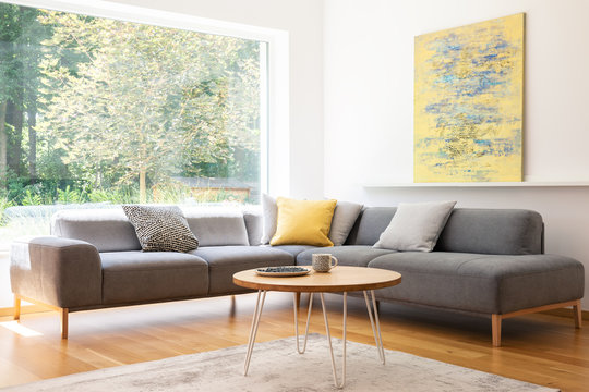 Cushions on grey corner couch in bright living room interior with yellow painting and table. Real photo